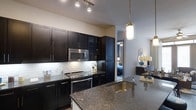 This modern single-floor residence showcases a thoughtfully designed open-concept layout that seamlessly connects the kitchen, dining, and living spaces. The kitchen serves as the heart of the home, featuring rich dark cabinetry that extends floor to ceiling, providing abundant storage. Striking granite countertops with deep, varied toning anchor the space, while a built-in undermount sink adds a refined touch. Stainless steel appliances, including a sleek electric range and over-the-range microwave, complement the contemporary aesthetic. A classic white subway tile backsplash offers a clean contrast against the dark cabinetry. Overhead, a combination of track lighting and elegant pendant lights illuminate the kitchen with warm, layered light. The open sightlines from the kitchen extend into the living and bedroom areas, enhancing the sense of spaciousness and connectivity throughout the residence. The overall ambiance is sophisticated and modern, with a carefully curated palette of dark, neutral, and metallic tones that lend the space a polished, upscale character well-suited for both everyday living and entertaining.