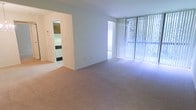 This single-floor unit features an open living area finished with wall-to-wall beige carpeting that lends a clean, neutral aesthetic throughout. The space is defined by a floor-to-ceiling sliding glass door fitted with vertical blinds, which allows generous natural light to fill the room and provides an outward view of surrounding greenery. The bright, white walls contribute to an airy and open atmosphere, enhancing the sense of spaciousness within the living area. An open passageway connects the living space to the kitchen, which is visible in the background and features tiled flooring, providing a practical contrast to the carpeted living area. The kitchen appears to include standard appliances and cabinetry. The layout flows naturally between the two areas, making the space feel cohesive and functional. Overall, the unit offers a straightforward, well-lit interior that balances comfort and practicality, with the large glazed door serving as a prominent architectural feature that anchors the living space with both light and outdoor connection.