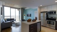 This contemporary single-floor space showcases a thoughtfully designed open-concept layout that seamlessly connects the kitchen and living areas. The kitchen is anchored by a sleek island with dark wood cabinetry and a crisp white countertop, offering both prep space and casual seating. Stainless steel appliances, including a full-sized refrigerator, complement the modern aesthetic, while white upper cabinetry provides ample storage. Light hardwood flooring flows throughout, adding warmth to the clean, contemporary palette. A bold teal accent wall in the living area serves as a striking focal point, framing a wall-mounted flat-screen television and adding a pop of color to the otherwise neutral tones. The living space is generously proportioned, furnished with a comfortable sofa and a low-profile coffee table atop a patterned area rug. Floor-to-ceiling windows flood the space with abundant natural light and offer sweeping panoramic city views, creating an airy, open ambiance. Exposed concrete columns and a designer pendant light fixture add industrial-chic character to the space, balancing the modern finishes with raw architectural detail.