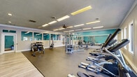 Located at 311 S Lasalle St in Durham, NC, this fitness facility is thoughtfully designed to accommodate a comprehensive workout experience across a single, open floor. The space is anchored by a generously sized main exercise room featuring durable rubber flooring, crown molding, and signature teal accent walls that lend the space a vibrant, energizing atmosphere. Large wall-spanning mirrors amplify the sense of openness while allowing users to monitor their form during exercise. The main gym area is outfitted with a diverse array of equipment, including treadmills, elliptical machines, rowing machines, cable weight systems, adjustable benches, and a dumbbell rack — catering to both cardio and strength training. A row of windows along one wall invites natural light and outdoor views, while flush panel lighting and ceiling fans ensure comfortable conditions throughout. Adjacent to the main floor, a dedicated studio room features warm hardwood flooring, recessed lighting, and its own set of mirrors. This room is equipped for more dynamic workouts, housing spin bikes, medicine balls, a Reflect interactive fitness mirror, and colorful free weights. A connecting hallway links the two areas, completing the functional layout of this well-rounded fitness destination.