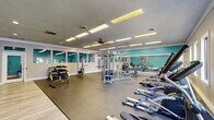 Located at 311 S Lasalle St in Durham, NC, this fitness facility is thoughtfully designed to accommodate a comprehensive workout experience across a single, open floor. The space is anchored by a generously sized main exercise room featuring durable rubber flooring, crown molding, and signature teal accent walls that lend the space a vibrant, energizing atmosphere. Large wall-spanning mirrors amplify the sense of openness while allowing users to monitor their form during exercise. The main gym area is outfitted with a diverse array of equipment, including treadmills, elliptical machines, rowing machines, cable weight systems, adjustable benches, and a dumbbell rack — catering to both cardio and strength training. A row of windows along one wall invites natural light and outdoor views, while flush panel lighting and ceiling fans ensure comfortable conditions throughout. Adjacent to the main floor, a dedicated studio room features warm hardwood flooring, recessed lighting, and its own set of mirrors. This room is equipped for more dynamic workouts, housing spin bikes, medicine balls, a Reflect interactive fitness mirror, and colorful free weights. A connecting hallway links the two areas, completing the functional layout of this well-rounded fitness destination.