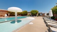 Located at 9380 Quadrangle St in Lorton, Virginia, this outdoor aquatic facility is a well-appointed community pool complex set against the backdrop of classic brick architecture. The grounds are thoughtfully designed to accommodate both active swimmers and leisure seekers alike.

At the heart of the facility is a generously sized lap pool with clearly marked lanes, complemented by a lifeguard station and convenient pool entry steps. Adjacent to it sits a shallower leisure pool featuring a distinctive mushroom-style water fountain, making it well-suited for casual recreation. Both pools are maintained in pristine condition with clear, inviting blue water.

The surrounding deck is spacious and finished in light-toned concrete, offering ample room for movement and relaxation. Rows of modern white lounge chairs with accompanying side tables and retractable umbrellas line the perimeter, providing comfortable spots to unwind in the sun or shade. Seating areas with tables and chairs are also available near the gated entrance for more social gatherings.

Lush landscaping frames the entire space, with mature trees, flowering shrubs, and colorful plantings adding vibrant greenery and natural privacy. A brick support building provides accessible restroom facilities and water fountains. The entire area is enclosed by a secure black iron fence, ensuring a safe and contained environment.