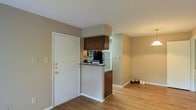 This single-floor unit offers a practical and welcoming layout that makes efficient use of its space. Upon entry, residents are greeted by warm hardwood flooring that extends throughout the main living area, lending a cohesive and inviting feel to the interior. The kitchen is positioned adjacent to the entryway and features a raised peninsula counter that serves as both a functional workspace and a visual divider between the kitchen and the living area. Dark wood cabinetry, including an overhead cabinet above the peninsula, provides ample storage while adding a contrasting design element against the neutral, beige-toned walls. The kitchen is equipped with essential appliances, including a microwave and a full-sized refrigerator, making it well-suited for everyday living. White trim and baseboards throughout the space offer a clean, classic finish that complements the warm tones of the flooring and cabinetry. The living area flows naturally from the kitchen, benefiting from an open feel and soft overhead lighting. The overall ambiance is calm and functional, with a neutral color palette that offers a versatile backdrop for a variety of interior styles. The single-floor design ensures easy accessibility and a straightforward, efficient use of the available space.