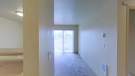 This single-floor space presents a clean, straightforward layout centered around a bright main living area. The room is finished with wall-to-wall carpet in a neutral gray tone, contributing to a calm and understated aesthetic throughout. Warm beige walls are consistent across the space, lending a cohesive and inviting atmosphere.

A prominent sliding glass door serves as the focal point of the main living area, fitted with vertical blinds and framing an outdoor view of surrounding buildings and hillside scenery. This large opening allows an abundance of natural light to filter into the space, creating a bright and airy feel even on overcast days. The door provides direct access to what appears to be a balcony or exterior patio area.

The ceiling height is modest and consistent, reinforcing a straightforward residential character. Light switch panels and electrical outlets are positioned along the walls at standard intervals. A wall-mounted heating vent is visible near floor level, indicating a built-in climate control system. The overall design is simple and functional, offering a neutral canvas well-suited to a variety of interior arrangements and personal styles.