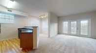 This single-floor residence offers a well-organized layout with distinct living, kitchen, bedroom, bathroom, and laundry areas. The open-concept living room is spacious and bright, featuring neutral-toned carpet, vaulted ceilings with track lighting, and a set of French doors that invite natural light and provide outdoor access. A kitchen peninsula with a dark countertop and undermount sink separates the kitchen from the living area, reinforcing the open feel.

The kitchen is a functional galley-style space with warm wood cabinetry, dark countertops, and black appliances including a range, refrigerator, dishwasher, and built-in microwave. Light hardwood flooring runs throughout the kitchen, complementing the cabinetry tones. Two windows fitted with blinds provide natural light.

The bedroom is generously sized with plush carpeting, a window overlooking the outdoors, and direct access to a walk-in closet equipped with wire shelving. The bathroom features light hardwood flooring, a white tile tub-and-shower combination, and standard fixtures. A dedicated laundry area houses a full-size side-by-side washer and dryer with overhead wire shelving for added storage.