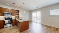 This well-appointed single-floor unit offers a thoughtfully designed open-concept layout that seamlessly connects the kitchen and living area. The kitchen is a standout feature, showcasing warm-toned wooden cabinetry, granite countertops, and a peninsula with an undermount sink that doubles as a breakfast bar. Stainless steel appliances — including a refrigerator, electric range, and over-the-range microwave — are complemented by a tile backsplash and built-in open shelving for added storage and display. Pendant lighting above the peninsula adds a stylish touch. Light hardwood-style flooring flows throughout the main living area, contributing to a bright and airy atmosphere. A large sliding glass door floods the living space with natural light and provides direct access to an outdoor area. The bedrooms are carpeted and finished with neutral tones, offering a calm and comfortable retreat. Each bedroom includes a closet with built-in shelving and rod for organized storage. The bathroom is clean and functional, featuring granite countertops, warm wood vanity cabinetry with brushed nickel hardware, a tub-shower combination, and tile flooring. The overall design maintains a consistent modern aesthetic throughout, with recessed lighting, clean white walls, and cohesive finishes.