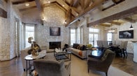 This single-story clubhouse is defined by its dramatic vaulted ceiling, where exposed wooden beams cross overhead in a rich, warm tone, complementing the natural stone construction throughout the space. The focal point of the main lounge area is a striking floor-to-ceiling fireplace clad in light-toned stacked stone, framed by a classic mantle and flanked by large windows dressed in patterned drapery that allow abundant natural light to fill the room. Recessed and track lighting along the beamed ceiling provides additional illumination, enhancing the inviting ambiance during evening hours. The lounge is furnished with a sophisticated arrangement of plush sofas and upholstered armchairs in neutral tones, centered around a sturdy rectangular coffee table featuring a decorative chess set. Side tables with accent lamps and carefully chosen décor add warmth and character throughout. Glass-paneled doors at the rear offer views to an outdoor area, reinforcing the connection between interior comfort and the surrounding environment. The overall atmosphere is refined yet welcoming, combining rustic architectural elements with polished furnishings to create a versatile and elegant communal gathering space.