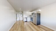 This single-floor space is thoughtfully designed to maximize functionality within a compact footprint. The open layout centers around a modern kitchen that serves as the focal point of the unit, outfitted with a full suite of stainless steel appliances including a refrigerator, electric range, over-the-range microwave, and dishwasher. Crisp white shaker-style cabinetry lines the walls above and below the counter, complemented by a clean white tile backsplash and a stainless steel sink. The light hardwood-style flooring runs continuously throughout the space, lending warmth and a sense of openness. Recessed lighting illuminates the interior, while a large window allows natural light to brighten the living area, offering views to the exterior. A wall-mounted mini-split air conditioning unit provides climate control, and a stacked washer and dryer unit is conveniently integrated into the space. Neutral white walls and clean-lined finishes give the interior a fresh, modern aesthetic. An electrical panel is accessible from the open living area, and multiple outlets are positioned throughout. The overall ambiance is bright, clean, and move-in ready, reflecting a practical and efficient design sensibility.