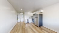 This single-floor space is thoughtfully designed to maximize functionality within a compact footprint. The open layout centers around a modern kitchen that serves as the focal point of the unit, outfitted with a full suite of stainless steel appliances including a refrigerator, electric range, over-the-range microwave, and dishwasher. Crisp white shaker-style cabinetry lines the walls above and below the counter, complemented by a clean white tile backsplash and a stainless steel sink. The light hardwood-style flooring runs continuously throughout the space, lending warmth and a sense of openness. Recessed lighting illuminates the interior, while a large window allows natural light to brighten the living area, offering views to the exterior. A wall-mounted mini-split air conditioning unit provides climate control, and a stacked washer and dryer unit is conveniently integrated into the space. Neutral white walls and clean-lined finishes give the interior a fresh, modern aesthetic. An electrical panel is accessible from the open living area, and multiple outlets are positioned throughout. The overall ambiance is bright, clean, and move-in ready, reflecting a practical and efficient design sensibility.