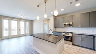 Located at 101 Cay Plaza in Savannah, Georgia, this well-appointed 2-bedroom, 2-bathroom residence offers a thoughtfully designed single-level floor plan with a clean, contemporary aesthetic. The open-concept kitchen and living area serves as the heart of the home, anchored by a central island with dark granite countertops and an undermount sink. Pendant lighting hangs above the island, complementing the recessed lights overhead. The kitchen is equipped with stainless steel appliances—including a gas range, built-in microwave, and French door refrigerator—alongside sleek gray cabinetry and a white subway tile backsplash. Light wood-look flooring flows seamlessly throughout the entire residence. The living area opens to a private balcony through glass French doors, bringing in generous natural light and outdoor views. Both bedrooms are comfortably sized, featuring large windows and ceiling fans. The primary bedroom includes a walk-in closet with wire shelving. Each bathroom is finished with gray cabinetry, dark granite countertops, and marble-style tile surrounds. The primary bath features a glass-enclosed shower, while the secondary bath offers a full tub-shower combination. Two hallways and additional closets provide ample storage throughout.