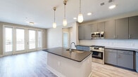 Located at 101 Cay Plaza in Savannah, Georgia, this well-appointed 2-bedroom, 2-bathroom residence offers a thoughtfully designed single-level floor plan with a clean, contemporary aesthetic. The open-concept kitchen and living area serves as the heart of the home, anchored by a central island with dark granite countertops and an undermount sink. Pendant lighting hangs above the island, complementing the recessed lights overhead. The kitchen is equipped with stainless steel appliances—including a gas range, built-in microwave, and French door refrigerator—alongside sleek gray cabinetry and a white subway tile backsplash. Light wood-look flooring flows seamlessly throughout the entire residence. The living area opens to a private balcony through glass French doors, bringing in generous natural light and outdoor views. Both bedrooms are comfortably sized, featuring large windows and ceiling fans. The primary bedroom includes a walk-in closet with wire shelving. Each bathroom is finished with gray cabinetry, dark granite countertops, and marble-style tile surrounds. The primary bath features a glass-enclosed shower, while the secondary bath offers a full tub-shower combination. Two hallways and additional closets provide ample storage throughout.