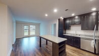 This single-floor residence offers a thoughtfully designed layout that balances style and functionality. The heart of the home is a modern kitchen outfitted with rich dark cabinetry, light granite countertops, and a full suite of stainless steel appliances, including a refrigerator, range, and microwave. A central island with matching granite provides additional prep space and open shelving below, while recessed lighting illuminates the hardwood-style flooring throughout. The kitchen flows seamlessly into an open living area bathed in natural light from large windows and glass doors that offer views of the surrounding landscape. The bedrooms feature neutral-toned carpet flooring and generous windows that invite plenty of daylight, creating bright and comfortable retreats. One bedroom includes access to a well-organized walk-in closet with wire shelving, positioned conveniently adjacent to a full bathroom. The bathroom is finished with a classic black-and-white mosaic tile floor, a clean white vanity with a modern faucet, and a full tub and shower combination. Throughout the residence, clean white walls, recessed lighting, and consistent finishes contribute to a cohesive, move-in-ready aesthetic.