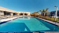 This outdoor amenity space centers around a generously sized swimming pool with sparkling blue water, framed by clean, light-colored pool decking that invites relaxation and leisure. The pool area is well-appointed with multiple lounge chairs and shade umbrellas, offering ample space for sunbathing and socializing throughout the day. A safety ring station is visibly positioned poolside, reflecting a commitment to guest well-being. The adjacent single-story clubhouse building features a contemporary, flat-roofed design with a neutral stucco exterior and large glass-paneled openings, providing a seamless transition between indoor and outdoor spaces. The facility is enclosed by a perimeter fence, ensuring privacy and security for its users. Mature palm trees and lush landscaping frame the surroundings, lending a tropical, resort-like ambiance to the environment. Under clear blue skies, the space benefits from abundant natural sunlight that reflects off the pool's surface, enhancing the inviting atmosphere. The overall setting conveys a well-maintained, modern community amenity designed for outdoor recreation and relaxation.