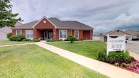 This single-story structure is built with classic red brick construction, giving it a timeless and sturdy appearance. The exterior showcases a prominent gabled roofline with a decorative circular window accent, adding architectural character to the facade. A covered entryway with an arched opening serves as the main point of entry, framed by neatly trimmed shrubs and ornamental plantings that enhance the building's curb appeal. Multiple traditional-style windows allow natural light to filter into the interior spaces. A wide concrete walkway leads directly from the street to the front entrance, providing clear and accessible access. The surrounding grounds are well-kept, featuring a lush, manicured lawn that extends across the front and sides of the property. A white fence is visible along the side of the building, delineating the outdoor space. The overall exterior presents a clean, professional, and inviting appearance, suggesting a well-maintained facility designed to be both functional and welcoming to visitors and occupants alike.