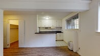 This single-floor space offers a practical and functional layout with clean, neutral finishes throughout. The kitchen is a standout area, featuring white upper and lower cabinetry providing ample storage, dark laminate countertops, a stainless steel sink, an electric range, and a full-size refrigerator. White tile flooring runs through the kitchen area, while the rest of the unit transitions to hardwood-style flooring that extends through the living and bedroom spaces. A half-wall peninsula separates the kitchen from the adjacent living area, creating an open, connected feel. The living area is notably bright, with a row of multiple windows along one wall that invite generous natural light and offer views of surrounding greenery. The bedroom is a calm, open space with light-toned walls and the same warm hardwood-style floors, bathed in soft, diffused sunlight. The bathroom includes a full-size soaking tub with a clean white surround panel system, chrome fixtures, and built-in wall shelving niches. Throughout the unit, walls are painted in soft, warm whites and creams, lending a clean and cohesive ambiance. The overall design is straightforward and move-in ready, ideal for comfortable everyday living.