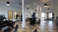 This fitness center is designed to accommodate a comprehensive workout experience within a spacious, single-floor layout. The space features a distinctive checkered floor pattern in neutral tones of beige and gray, lending a polished, professional look throughout. Sturdy white structural columns punctuate the open floor plan, complementing the crisp white walls and creating a clean, airy atmosphere. Overhead, ceiling fans and pendant black industrial-style light fixtures provide both comfort and focused illumination across the workout areas. Natural light filters in through windows along the perimeter, enhancing the bright and energizing environment. The facility is outfitted with a wide selection of strength training machines, including multi-function cable and weight machines, as well as benches and free-weight stations. Cardio equipment is also present, ensuring a well-rounded fitness offering. Large mirrors are mounted along the walls, amplifying the sense of space and allowing users to monitor their form during exercises. The overall ambiance is modern and functional, with a thoughtful layout that allows for comfortable movement between equipment stations.
