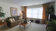 This single-floor residence offers a comfortable and well-appointed living environment designed with a classic, understated aesthetic. The living room serves as the heart of the space, featuring plush wall-to-wall carpeting that adds warmth underfoot and contributes to the room's cozy atmosphere. Large windows dressed with warm golden curtains and horizontal blinds allow an abundance of natural light to fill the room, creating a bright and welcoming ambiance. The neutral wall tones provide a versatile backdrop that complements the earthy, muted furnishings throughout. A generously sized sofa dressed with decorative pillows anchors the seating area, paired with a mid-century-style wooden coffee table that adds a touch of timeless charm. Decorative accents, including a framed landscape artwork on the wall, a floor lamp, and tasteful greenery on the coffee table, lend the space a lived-in, homey character. Additional decorative pieces, such as a small trunk and a wooden side table, round out the room's carefully considered layout. The overall ambiance is calm, warm, and inviting — a space that balances functionality with a comfortable residential character well-suited for everyday living.