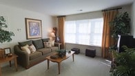 This single-floor residence offers a comfortable and well-appointed living environment designed with a classic, understated aesthetic. The living room serves as the heart of the space, featuring plush wall-to-wall carpeting that adds warmth underfoot and contributes to the room's cozy atmosphere. Large windows dressed with warm golden curtains and horizontal blinds allow an abundance of natural light to fill the room, creating a bright and welcoming ambiance. The neutral wall tones provide a versatile backdrop that complements the earthy, muted furnishings throughout. A generously sized sofa dressed with decorative pillows anchors the seating area, paired with a mid-century-style wooden coffee table that adds a touch of timeless charm. Decorative accents, including a framed landscape artwork on the wall, a floor lamp, and tasteful greenery on the coffee table, lend the space a lived-in, homey character. Additional decorative pieces, such as a small trunk and a wooden side table, round out the room's carefully considered layout. The overall ambiance is calm, warm, and inviting — a space that balances functionality with a comfortable residential character well-suited for everyday living.