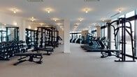 This expansive, single-level fitness facility is designed to accommodate a comprehensive range of training needs within a clean, contemporary aesthetic. The space is clearly divided into distinct functional zones, each thoughtfully equipped and finished.

The primary strength training area features light-toned carpeting underfoot and is anchored by multiple power racks and squat stations loaded with Olympic barbells and weight plates. Adjustable benches are positioned nearby, and a full dumbbell rack lines one mirrored wall, amplifying the sense of depth and scale throughout this zone.

The cardio section runs along the perimeter near the floor-to-ceiling windows, housing a comprehensive lineup of Matrix-brand treadmills, elliptical trainers, stair climbers, and cable machines. The generous glazing in this area floods the space with natural light and provides open outdoor views.

A separate studio area distinguished by wide-plank hardwood flooring offers a more open, flexible training environment. This room features a large wall-mounted mirror, a built-in wooden shelving unit stocked with medicine balls, stability balls, and yoga mats, along with spin bikes positioned near the expansive glass doors leading outside.

Throughout, the facility maintains a bright, neutral palette with recessed and flush-mount lighting, white walls, and structural columns that define zones without obstructing the overall open feel.