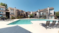 Located on S 5th Street in Saint Charles, Missouri, this outdoor pool area serves as a central amenity for a multi-building residential community. The space is defined by a generously sized rectangular swimming pool with clear blue water, encircled by a broad concrete sun deck that provides ample room for relaxation and recreation. Multiple wrought-iron lounge chairs and seating arrangements are positioned throughout the deck, offering residents comfortable spots to enjoy the outdoors under open skies.

The pool area is enclosed by a black metal fence with a designated entrance, and clearly posted signage ensures the safety of all users. A shaded seating area with a blue umbrella and additional chairs provides a cooler retreat alongside the main deck. The surrounding multi-story residential buildings feature traditional architecture with neutral-toned siding, pitched rooflines, prominent chimneys, and private balconies that overlook the pool courtyard, creating an enclosed and community-oriented atmosphere.

Practical amenities include exterior wall-mounted lighting fixtures and direct building access via glass-panel doors. Well-maintained hedges and mature trees add a touch of greenery to the hardscaped environment. The overall setting is bright, open, and inviting, ideal for warm-weather outdoor enjoyment in a well-established residential community.