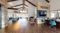Located at 1430 Bell Rd in Nashville, TN, the Whetstone Flats clubhouse is a generously sized, single-floor community space that blends rustic and contemporary design elements throughout. The centerpiece of the space is a dramatic vaulted ceiling with exposed wooden beams, anchoring an expansive open-plan layout finished with rich hardwood-style flooring. A reclaimed wood accent wall prominently displays the Whetstone Flats branding alongside a wall-mounted TV and a sleek built-in fireplace, creating a warm and welcoming focal point upon entry. The leasing area features wooden desks with teal accent chairs and glass-partitioned private offices accessible via sliding barn doors, maintaining an open yet professional atmosphere. A comfortable lounge area offers multiple seating arrangements, including a cozy sitting nook with a fireplace framed in textured tile, built-in geometric shelving, and curated artwork. A dedicated meeting room with a teal ceiling, linear pendant lighting, and upholstered seating provides a refined space for gatherings. The casual dining area, outfitted with teal chairs and a playful mosaic feature wall, opens directly to an outdoor pool area through large glass doors, filling the space with abundant natural light.
