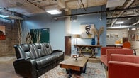 This expansive single-floor space masterfully blends raw industrial character with curated interior design. Throughout the layout, exposed concrete ceilings reveal an intricate network of pipes, ducts, and structural beams, while polished concrete floors and original brick walls reinforce the building's historic industrial roots. Distinctive cast-iron columns with weathered patinas anchor the open floor plan, serving as striking architectural focal points.

The space is organized across several distinct zones. A well-appointed lounge area features a large leather reclining sofa, upholstered armchairs in warm terracotta tones, and a decorative area rug, all set against soft blue-painted walls adorned with curated artwork. A circular-base console shelf and accent lamps add refined detail to this relaxed gathering area.

An adjacent open area provides generous flexibility, with seating vignettes positioned near brick accent walls and a vintage-style elevator with heavy steel doors. Further in, a dedicated room with barrel-vaulted concrete ceilings houses a poker or game table, lending an intimate, cave-like atmosphere. A separate zone includes a bar-height counter with stools and cabinetry, suitable for catering or social functions. The combination of raw architectural elements and thoughtful furnishings creates a dynamic, versatile environment throughout.