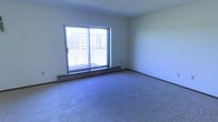 This single-floor space features a generously sized main room finished with wall-to-wall carpet in a neutral blue-gray tone, providing a practical and comfortable foundation underfoot. The room is brightened by a large sliding glass door that fills the space with natural light and opens directly onto a private balcony, framed by a simple metal railing. Views through the glass look out toward an adjacent multi-story building, offering an urban backdrop. A baseboard heating unit runs beneath the sliding door, providing an efficient source of warmth along the exterior wall. The walls are painted a clean, crisp white, complementing the light-filled atmosphere, while the textured ceiling adds a subtle visual detail overhead. The overall layout is open and unobstructed, allowing for flexible furniture arrangements and a versatile use of the space. The combination of natural light, outdoor access, and a neutral interior palette creates a straightforward, functional environment well-suited for a variety of living arrangements.
