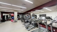 This fitness center offers a thoroughly equipped exercise environment designed to support a wide range of workout routines. The space is laid out across a single floor, maximizing accessibility and ease of movement between stations. A row of cardio machines — including ellipticals, treadmills, and stationary bikes — lines one side of the room, each unit equipped with individual monitor screens for entertainment during workouts. The strength training area features a dedicated cable and weight machine zone, while a separate section houses stability balls and additional functional fitness equipment. Large floor-to-ceiling mirrors run along the walls, amplifying the sense of space and allowing users to monitor their form. The interior design incorporates deep burgundy accent panels against neutral tones, giving the space a polished, professional atmosphere. Bright, recessed linear lighting illuminates the entire floor evenly, ensuring excellent visibility throughout. The flooring is a smooth, durable surface well-suited for both machine-based and functional training. Overall, the fitness center is thoughtfully organized to accommodate multiple users simultaneously while maintaining a clean, uncluttered environment conducive to focused exercise.