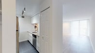 This single-floor unit offers a clean, modern aesthetic throughout, defined by a consistent white color palette, light-toned hardwood flooring in the living areas, and an abundance of natural light. The open living room is a standout feature, with floor-to-ceiling vertical blinds lining an expansive window wall that floods the space with daylight and creates an airy, open ambiance. Track lighting is installed throughout, providing flexible and well-distributed illumination across all areas. The kitchen is thoughtfully designed with crisp white cabinetry extending to the ceiling for ample storage, light quartz countertops, a white subway tile backsplash, and a full suite of stainless steel appliances including an electric range, over-the-range microwave, dishwasher, and sink with a modern gooseneck faucet. Dark tile flooring in the kitchen provides a subtle contrast to the surrounding light finishes. A stacked washer and dryer unit is conveniently tucked within the kitchen area. The bathroom continues the space's clean design language, featuring a white floating vanity with an undermount sink, a large mirror, hexagonal tile flooring, and a full tub-shower combination with white tile surround. Overall, the space presents a polished, move-in-ready interior with a cohesive minimalist character.