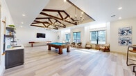 This expansive single-floor amenity center spans a generously sized footprint and is thoughtfully organized into four distinct functional areas connected by a bright, art-lined corridor with light hardwood flooring throughout. The game room serves as the architectural centerpiece, featuring soaring vaulted ceilings adorned with dark wood exposed beam trusses and dual chandelier lighting. A full-size billiards table and shuffleboard table anchor the space, complemented by a built-in wet bar with dark cabinetry, open shelving, and a microwave. Large windows fill the room with natural light and exterior views. Adjacent to the game room, a lounge area offers a comfortable seating arrangement with a sectional sofa, additional accent chairs, and windows overlooking the surroundings. The fully equipped kitchen features stainless steel appliances, dark cabinetry, a subway tile backsplash, open shelving, and a large island with seating. A dedicated fitness center contrasts the lighter common areas with dark walls, vaulted ceilings, rubber flooring, floor-to-ceiling mirrors, and a comprehensive array of cardio and strength-training equipment, including treadmills, stationary bikes, and a cable machine system.
