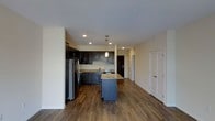 This well-appointed single-floor residence offers a thoughtfully designed open-concept layout that seamlessly connects the kitchen and living areas. The kitchen is a standout feature, showcasing rich dark cabinetry with detailed raised-panel doors, granite countertops, and stainless steel appliances including a full-size refrigerator. A central kitchen island with matching granite provides additional prep space and casual seating, while a stylish pendant light above adds a warm, focused accent. Recessed lighting throughout ensures the space is bright and evenly lit. Durable hardwood-style flooring in a warm medium-brown tone runs throughout, adding warmth and a sense of continuity across the open floor plan. The neutral, light-toned walls complement the darker cabinetry and flooring, creating a balanced and inviting aesthetic. Additional doors visible from the main living area suggest well-placed access to further rooms or storage, contributing to a practical and functional layout. Overall, the space combines modern finishes with a clean, open design that is both stylish and highly livable.