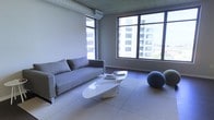 This single-floor space embodies a clean, modern aesthetic with a distinctly minimalist sensibility. The main living area is anchored by large, floor-to-ceiling windows that flood the room with natural light and offer sweeping views of the surrounding urban skyline, creating a strong connection between the interior and the city beyond.

The space features neutral-toned walls and light-colored flooring that enhance the sense of openness and airiness throughout. A sleek gray sofa serves as the focal point of the seating area, complemented by a sculptural white coffee table with an organic, flowing form that adds an artistic touch to the room. Accent pieces, including a rounded, textured blue decorative ball, contribute subtle personality to the otherwise understated palette.

Exposed ductwork along the ceiling lends an industrial character to the space, balancing the refined furnishings with a raw, urban edge. The open layout allows for flexible use of the area, making it well-suited for both relaxing and entertaining. The overall ambiance is calm, sophisticated, and contemporary — a well-considered space that prioritizes light, simplicity, and thoughtful design details throughout.
