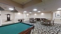 This single-floor recreational lounge is thoughtfully designed to provide a comfortable and engaging environment for leisure activities and social interaction. The centerpiece of the space is a full-size billiards table finished in rich mahogany with a classic green felt surface, complete with a pendant light overhead that highlights the table with focused illumination. Surrounding the billiards area, a boldly patterned carpet in swirling tones of grey, black, and cream adds a sense of energy and visual interest to the room. Multiple seating arrangements are distributed throughout, including upholstered chairs with traditional detailing and smaller round tables ideal for card games or casual conversation. The walls feature classic wainscoting and neutral cream tones that lend an air of refined elegance to the space. Recessed and flush-panel ceiling lights provide bright, even illumination throughout, complemented by a drop-panel ceiling with subtle architectural banding. Framed artwork accents the walls, adding a polished finishing touch. The overall ambiance is one of relaxed sophistication, making this lounge a welcoming destination for recreational and social activities.