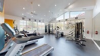 This fitness center offers a thoughtfully designed workout environment that combines practical functionality with an open, airy atmosphere. The space is anchored by soaring vaulted ceilings and flanked by large multi-pane windows on multiple walls, flooding the interior with natural light and providing views of the surrounding greenery. Ceiling fans are mounted overhead to ensure comfortable air circulation throughout the room.

The rubber flooring provides a durable, slip-resistant surface suitable for a wide range of exercise activities. Cardio equipment is prominently featured, including multiple Matrix treadmills and an elliptical trainer. Strength training is well-represented with a multi-station cable machine, weight benches, dumbbells, and stability balls. A dedicated spin bike and rowing equipment round out the diverse offering.

Full-length mirrors are installed along one wall, enhancing the sense of space while allowing users to monitor their form. A bold accent wall in vivid orange adds an energizing visual element, complemented by a wall-mounted flat-screen television for entertainment during workouts. Framed artwork is displayed throughout, contributing to a polished, welcoming atmosphere. Overall, the space is designed to support a comprehensive fitness routine in a bright and motivating environment.
