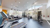 This fitness center offers a thoughtfully designed workout environment that combines practical functionality with an open, airy atmosphere. The space is anchored by soaring vaulted ceilings and flanked by large multi-pane windows on multiple walls, flooding the interior with natural light and providing views of the surrounding greenery. Ceiling fans are mounted overhead to ensure comfortable air circulation throughout the room.

The rubber flooring provides a durable, slip-resistant surface suitable for a wide range of exercise activities. Cardio equipment is prominently featured, including multiple Matrix treadmills and an elliptical trainer. Strength training is well-represented with a multi-station cable machine, weight benches, dumbbells, and stability balls. A dedicated spin bike and rowing equipment round out the diverse offering.

Full-length mirrors are installed along one wall, enhancing the sense of space while allowing users to monitor their form. A bold accent wall in vivid orange adds an energizing visual element, complemented by a wall-mounted flat-screen television for entertainment during workouts. Framed artwork is displayed throughout, contributing to a polished, welcoming atmosphere. Overall, the space is designed to support a comprehensive fitness routine in a bright and motivating environment.