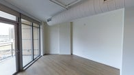 This single-floor space showcases a clean, modern industrial aesthetic that balances functionality with contemporary design. The open layout provides a versatile blank canvas, with smooth white walls offering ample opportunity for personalization or commercial use.

A standout architectural feature is the large, exposed cylindrical ductwork that runs prominently across the ceiling, lending an industrial character that complements the otherwise minimalist interior. The ceiling itself is finished in white, maintaining a bright and airy atmosphere throughout.

The flooring consists of warm hardwood-style planks that run the length of the space, adding a natural, grounding element that contrasts tastefully with the crisp white walls and industrial overhead details.

Floor-to-ceiling glazed panels along one wall flood the interior with generous natural light, creating a seamless visual connection to the exterior. The expansive windows enhance the sense of openness while providing excellent daylighting throughout the day.

The space also includes a wall-mounted HVAC unit, ensuring climate comfort year-round. Electrical outlets are conveniently positioned along the walls, supporting a range of potential configurations and uses.