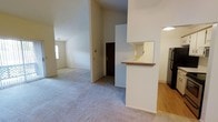 This single-story space offers a thoughtfully arranged floor plan that balances open living with defined functional areas. The interior is characterized by notably high ceilings that create a sense of openness and airiness throughout the main living zones. Neutral-toned walls and light carpeting run through the primary living areas, contributing to a clean, understated aesthetic.

A central entryway connects the main living spaces, with a pass-through kitchen counter serving as a natural divider between the kitchen and adjoining areas. The kitchen features wood-look flooring, white cabinetry, and a wrap-around counter with bar-style overhang, making it both practical and suitable for casual dining. A stainless steel refrigerator is visible from the main corridor.

The living areas are generously proportioned, with ceiling fans in multiple rooms aiding in airflow and comfort. A fireplace anchors one of the primary living spaces, adding a focal point and warmth to the room. Additional rooms branch off from the central corridor, providing separated spaces suitable for various uses. Natural light filters in through windows positioned throughout the layout, brightening the carpeted interiors and enhancing the overall welcoming ambiance of the space.
