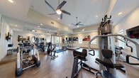 Located at 3544 Saint Johns Bluff Rd S in Jacksonville, Florida, this fitness center offers a clean, well-organized exercise space designed to support a comprehensive workout routine. The room features soaring ceilings that lend an open, airy feel, complemented by recessed lighting and dual ceiling fans that ensure comfortable airflow throughout. Durable, textured flooring covers the entire space, providing both traction and cushioning ideal for physical activity. Large wall-mounted mirrors visually expand the room and allow users to monitor their form during exercise. Natural light filters in through several windows fitted with zebra-style shades, offering a pleasant connection to the outdoors. The fitness center is well-stocked with a variety of equipment, including treadmills, an elliptical, a stationary bike, a multi-station cable and weight machine, a dumbbell rack with adjustable bench, and additional strength training apparatus. A wall-mounted flat-screen television provides entertainment during workouts. Motivational fitness artwork decorates the walls, adding energy to the environment. Glass doors at one end of the room open to the exterior, further enhancing ventilation and accessibility. The overall ambiance is bright, functional, and inviting, making this fitness center a practical and well-appointed amenity.