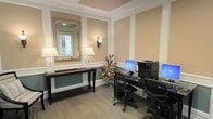 This single-floor space functions as a well-appointed business center, thoughtfully designed to blend functionality with classic elegance. The room features warm beige walls accentuated by crisp white wainscoting and decorative panel molding, lending the space a polished, formal aesthetic. A tray ceiling with crown molding adds architectural interest overhead, while textured carpeting grounds the room with a neutral, professional tone.

The workstation area is equipped with dual computer setups, each accompanied by an ergonomic task chair and a shared printer, providing all the essentials for productivity. Adjacent to the workstations, a sleek dark wood console table serves as a welcoming focal point, topped with matching glass-base table lamps and adorned with a vibrant floral arrangement in warm tones. Above the console, a large ornate gold-framed mirror reflects light and amplifies the sense of space.

A decorative wrought iron wall sconce is mounted between the paneled sections, contributing to the warm, ambient lighting throughout. The overall ambiance strikes a balance between a professional work environment and an inviting, upscale interior — ideal for guests or residents seeking a comfortable and well-equipped space to handle business needs.