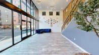 This contemporary building lobby combines industrial and modern design elements to create a welcoming, open-feel entrance. The space is anchored by dramatic floor-to-ceiling glass walls along the front facade, flooding the interior with abundant natural light and offering clear views of the surrounding streetscape. Large-format tile flooring in a neutral stone finish runs throughout, lending a clean, cohesive look underfoot.

A striking exposed brick wall adds warmth and character, contrasting beautifully with crisp white and slate-blue painted surfaces. The staircase is a standout architectural feature, rising prominently from the lobby floor with a wood handrail and sleek black metal balusters, framed against the bold brick backdrop. High ceilings throughout amplify the sense of volume and grandeur, while recessed lighting and contemporary ring-style pendant fixtures provide layered illumination.

A designated mailroom area lines one wall, outfitted with an extensive bank of individual mailboxes, offering residents convenient package and mail access directly from the lobby. Curated wall art, a curved navy blue sofa, and a potted olive tree introduce personality and comfort to the space. Together, these elements create a polished, inviting lobby that blends functionality with refined, modern aesthetics.