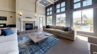 This single-floor community clubhouse or leasing center is defined by its impressive architectural design and upscale finishes throughout. The entry foyer immediately establishes a grand first impression with its distinctive slate tile flooring in warm earth tones, flanked by tall, classically styled white columns that frame the space with elegance. A coffered ceiling with blue-painted recessed panels and ornate lantern-style pendant fixtures draws the eye upward, emphasizing the generous ceiling height.

The central lounge area features a built-in console table beneath an expansive bank of multi-pane windows, which flood the interior with abundant natural light. A secondary lounge space showcases a dramatic two-story window wall offering direct views of an outdoor pool and patio area with lounge chairs and umbrellas, creating a seamless indoor-outdoor connection. This room is anchored by a stone-surround fireplace and furnished with large, plush neutral sofas layered with bold accent pillows atop rich area rugs.

Additional seating areas provide generous room for gathering, each benefiting from the soaring ceiling height reinforced by stacked transom windows. A dedicated office area is visible off the entry corridor, offering a functional workspace. The overall ambiance blends contemporary comfort with classic architectural detailing, resulting in a polished, welcoming environment throughout.