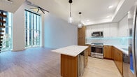 Located in the heart of downtown Houston, this contemporary two-story residence offers a thoughtfully designed layout across two generously proportioned floors. The open-concept main level combines the kitchen and living area into one cohesive, light-filled space. The kitchen is a standout feature, showcasing two-tone cabinetry with warm wood lower cabinets and crisp white upper cabinets, a marble-look tile backsplash with LED under-cabinet lighting, stainless steel appliances including a gas range, and a large central island topped with a quartz countertop and pendant lighting above. Light hardwood flooring and floor-to-ceiling windows flood the main level with natural light and frame striking views of the downtown Houston skyline and streetscape. A sleek staircase with a wood handrail and black cable railing connects to the upper level. The primary bedroom is a calm retreat featuring plush carpeting, a ceiling fan, generous natural light, and a barn door leading to a spacious walk-in closet with built-in shelving and organization systems. The primary bathroom is refined and spa-like, offering dual undermount sinks, a long wood-toned vanity, white quartz countertops, and backlit LED mirrors. A second full bathroom features a soaking tub with marble-look tile surround. Throughout, black hardware accents and a clean, minimalist palette create a cohesive modern aesthetic.