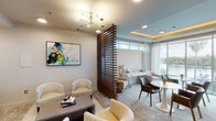 Located along the scenic Boardwalk Trail in Austin, TX, this co-working space delivers a polished, amenity-rich environment designed to accommodate both focused work and collaborative interaction. The single-floor layout is thoughtfully organized into distinct functional zones, separated by a striking slatted wood partition that adds architectural warmth while maintaining an open, airy feel throughout.

The lounge area features plush cream-toned armchairs, a low-profile coffee table, and a bold abstract artwork, anchored by a mid-century-inspired brass and glass chandelier overhead. Adjacent to this seating zone, a dedicated workstation area provides computer terminals, a printer, and ergonomic seating, accessible through a glass-etched entry marked "Co-Work."

A banquette seating section offers a channel-tufted upholstered bench paired with round tulip-style tables, ideal for informal meetings or individual work. The space transitions into a brighter lounge area crowned by a coffered ceiling with warm ambient LED lighting, where floor-to-ceiling windows frame sweeping views of the adjacent waterway and lush tree canopy. Natural light fills this zone generously, complemented by sheer drapery and a refined mix of contemporary furnishings that complete the sophisticated, welcoming atmosphere.