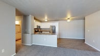 This single-floor residence features an open-concept layout that seamlessly connects the kitchen and living area, creating a cohesive and functional living environment. The kitchen is well-appointed with crisp white cabinetry offering ample storage, granite countertops with warm tonal variation, and a full suite of stainless steel appliances including a refrigerator, microwave, and range. A peninsula counter extends outward, providing a convenient breakfast bar area while visually defining the kitchen space without fully closing it off from the rest of the unit.

The transition from kitchen to living area is marked by a shift in flooring — wood-look hard surface flooring anchors the kitchen zone, while plush carpeting extends throughout the living area, adding warmth and comfort underfoot. The living space is generously proportioned, offering flexibility for a variety of furniture arrangements. Neutral gray walls and white trim provide a clean, understated backdrop throughout. Flush-mount ceiling light fixtures illuminate both areas, maintaining a consistent and practical lighting scheme. The overall ambiance is modern and move-in ready, with a clean, neutral palette that suits a range of personal styles.