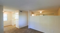 This single-story space presents a straightforward, functional layout with clearly defined areas separated by partial partition walls. The interior is finished in neutral beige tones throughout, contributing to a bright and cohesive aesthetic. Carpeted flooring covers the main living areas, adding warmth underfoot, while the textured ceilings and HVAC vents indicate a practical, well-maintained build.

A glimpse into the kitchen area reveals white cabinetry, a countertop workspace, and a window that allows natural light to filter in. The kitchen's light-filled character contrasts gently with the carpeted living zones adjacent to it. A half-wall partition separates portions of the space, creating a sense of division without fully closing off areas from one another.

A ceiling fan fixture is visible in one section, providing both lighting and ventilation. Additional ventilation grilles are positioned in the ceiling, suggesting a central air system. The overall layout feels open yet organized, with each zone transitioning naturally into the next. The clean walls and neutral palette make the space highly adaptable for a variety of uses and furnishing styles.