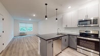 This single-floor unit offers a clean, contemporary design highlighted by light hardwood-style flooring and a crisp white color palette throughout. The open-concept kitchen flows seamlessly into the living area, creating a bright and functional shared space. The kitchen is outfitted with two-tone cabinetry — white upper cabinets paired with grey lower cabinets — complemented by a white subway tile backsplash, dark countertops, and a full suite of stainless steel appliances including a refrigerator, range, over-the-range microwave, and dishwasher. Stylish pendant lighting over the kitchen peninsula adds a modern accent. The living area receives natural light through windows with views of lush greenery, fitted with roller shades for privacy. Each bedroom is generously sized, featuring the same hardwood-style flooring, neutral walls, and individual climate control units positioned beneath the windows. The primary bathroom features floor-to-ceiling grey tile, a glass-enclosed walk-in shower, and a white vanity with modern fixtures. A second bathroom is designed with accessibility in mind, featuring large-format tile flooring, neutral wall paneling, and grab bars. The overall ambiance is modern, clean, and move-in ready.