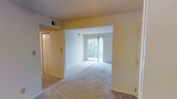 This single-floor residential space offers a clean, open layout designed for comfortable everyday living. The main living area is finished with soft, neutral-toned carpeting that extends throughout, creating a cohesive and welcoming atmosphere. Warm, light yellow accent walls contrast gently with the crisp white ceilings and trim, giving the interior a bright and airy character.

A standout feature of the living area is the full-width sliding glass door at the far end, which floods the room with abundant natural light and provides views of lush, mature greenery outside. This connection to the outdoors enhances the overall sense of openness and tranquility within the space.

The layout flows naturally between areas, with a wide open passageway connecting the main living zone to an adjacent room, allowing light to travel freely throughout. A closet with sliding doors is conveniently located near the entryway, offering practical storage. Visible HVAC vents in the ceiling indicate a central climate control system throughout.

Overall, the space presents a straightforward, functional design with neutral finishes that provide a versatile canvas, making it well-suited for a variety of furnishing styles and personal tastes.