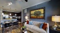 This single-floor residence showcases a thoughtfully designed open-concept layout that seamlessly connects the living and kitchen areas, creating a fluid and functional living environment. The living room draws immediate attention with its bold, slate-blue accent wall adorned with a large, ornately framed landscape painting that serves as a striking focal point. A tufted cream sofa, accented with rust and blue throw pillows, anchors the space, while warm hardwood flooring runs throughout, adding richness and continuity. A ceiling fan with track lighting overhead provides both comfort and ambiance. The kitchen is finished with dark espresso cabinetry, granite countertops, and a tiled backsplash, offering a sophisticated contrast to the lighter living area. Pendant lighting hangs above the kitchen island, casting a warm glow over the space. A dining area with modern seating sits at the intersection of the two spaces, reinforcing the open and connected feel of the floor plan. Crown molding details and decorative columns add architectural character throughout. The overall ambiance is modern yet inviting, with a carefully curated blend of textures, colors, and finishes that create a polished and comfortable interior.