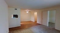 This single-story space offers a straightforward, functional layout with a blend of flooring materials that visually define different areas. The main living area features warm hardwood flooring that extends toward an arched passageway leading to an adjacent room, while a carpeted section in the foreground provides a softer, more comfortable zone — ideal for a seating or lounge area.

The ceiling is standard height and finished in a clean, neutral white, complemented by light beige walls throughout, creating a bright and open atmosphere. A ceiling fan with an integrated light fixture is centrally positioned, providing both air circulation and ambient lighting to the space.

A notable architectural feature is the pass-through window cut into the interior wall, which connects the living area to an adjoining space — likely a kitchen — allowing for easy communication and light transfer between rooms. A television is visible through the opening, suggesting versatile use of the connected space.

A doorway on the right side of the room and an arched opening at the rear provide access to additional areas of the home. HVAC vents are present on both the wall and ceiling, ensuring climate control throughout. The overall ambiance is clean, neutral, and move-in ready.