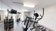 Located at 1722 N Walnut St in Bloomington, Indiana, this fitness room offers a well-organized exercise space designed to support a well-rounded workout routine. The room features durable rubber flooring throughout, providing a safe and practical surface for exercise. Bright flush-mounted ceiling fixtures ensure consistent, even lighting across the space.

The room is outfitted with a variety of equipment, including elliptical machines, a treadmill, and a multi-functional cable machine for strength training. A rack of dumbbells provides options for free weight exercises, while a water cooler and hand sanitizer station are conveniently positioned for user comfort and hygiene. A wall-mounted flat-screen television offers entertainment during workouts.

Full-length mirrors line one wall, visually expanding the room and allowing users to monitor their form. A large window with vertical blinds faces a green, wooded outdoor view, bringing in natural light and creating a pleasant exercise environment. A wall-mounted HVAC unit ensures climate control year-round. The clean, neutral white walls keep the atmosphere bright and focused. This fitness room delivers a comprehensive range of equipment and amenities within a compact, efficiently utilized footprint.